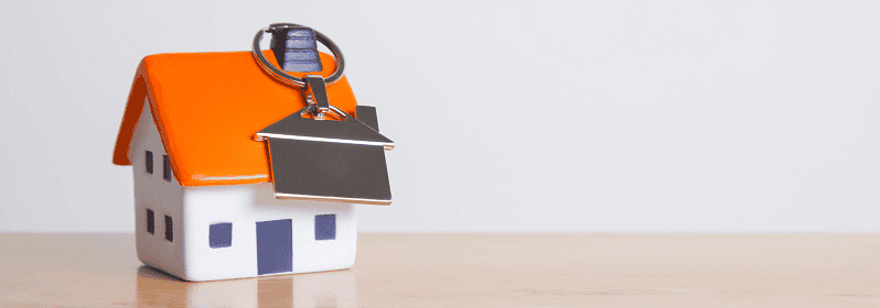A small model house on a wooden table with a key, representing securing a property investment and the concept of homeownership.
