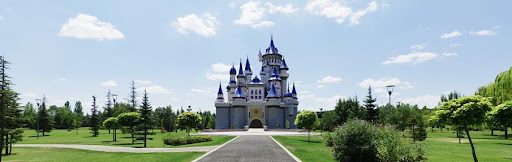 The iconic Fairytale Castle (Masal Şatosu) in Eskişehir's Sazova Park, seen at the end of a long central pathway, with its towering blue-topped spires set against a sunny blue sky with white clouds and surrounded by lush green parkland.