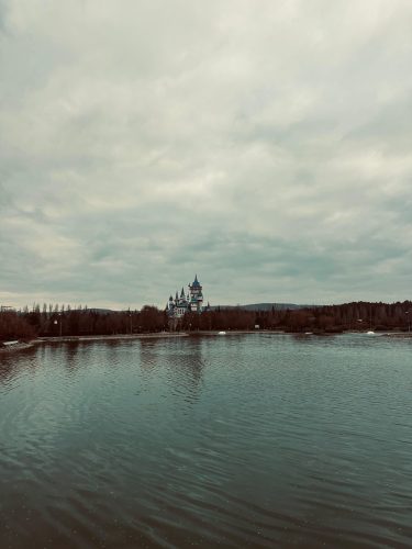A wide-angle landscape view of the Fairy Tale Castle (Masal Şatosu) in Eskişehir, sitting across a large, dark rippled lake under a dramatic cloudy sky.