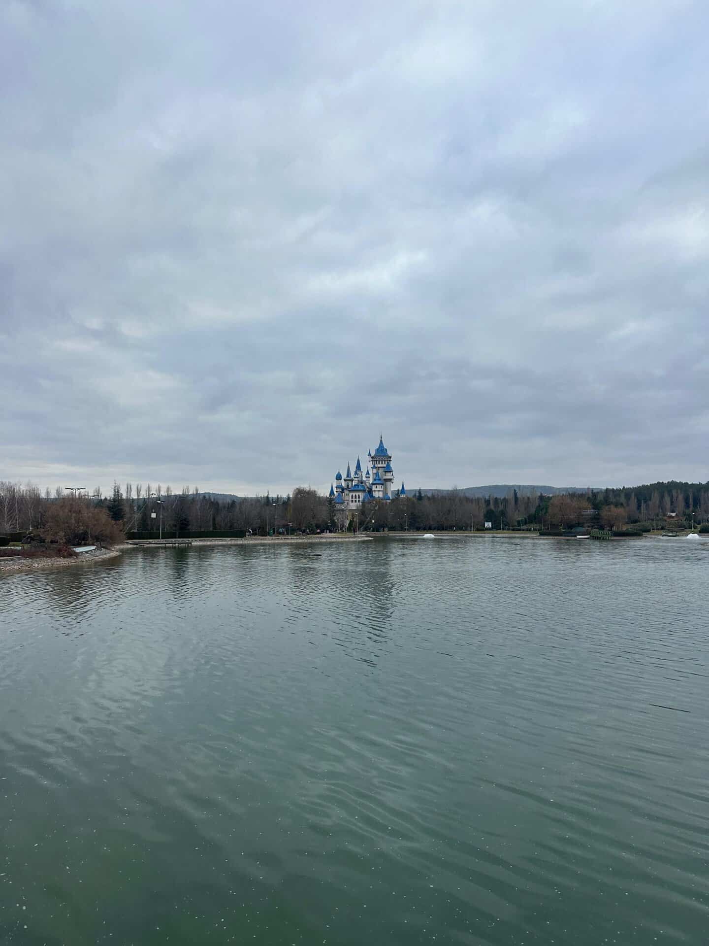 A wide landscape view of the blue-spired Fairy Tale Castle (Masal Şatosu) in Sazova Park, Eskişehir, seen from across a large lake under a cloudy sky.